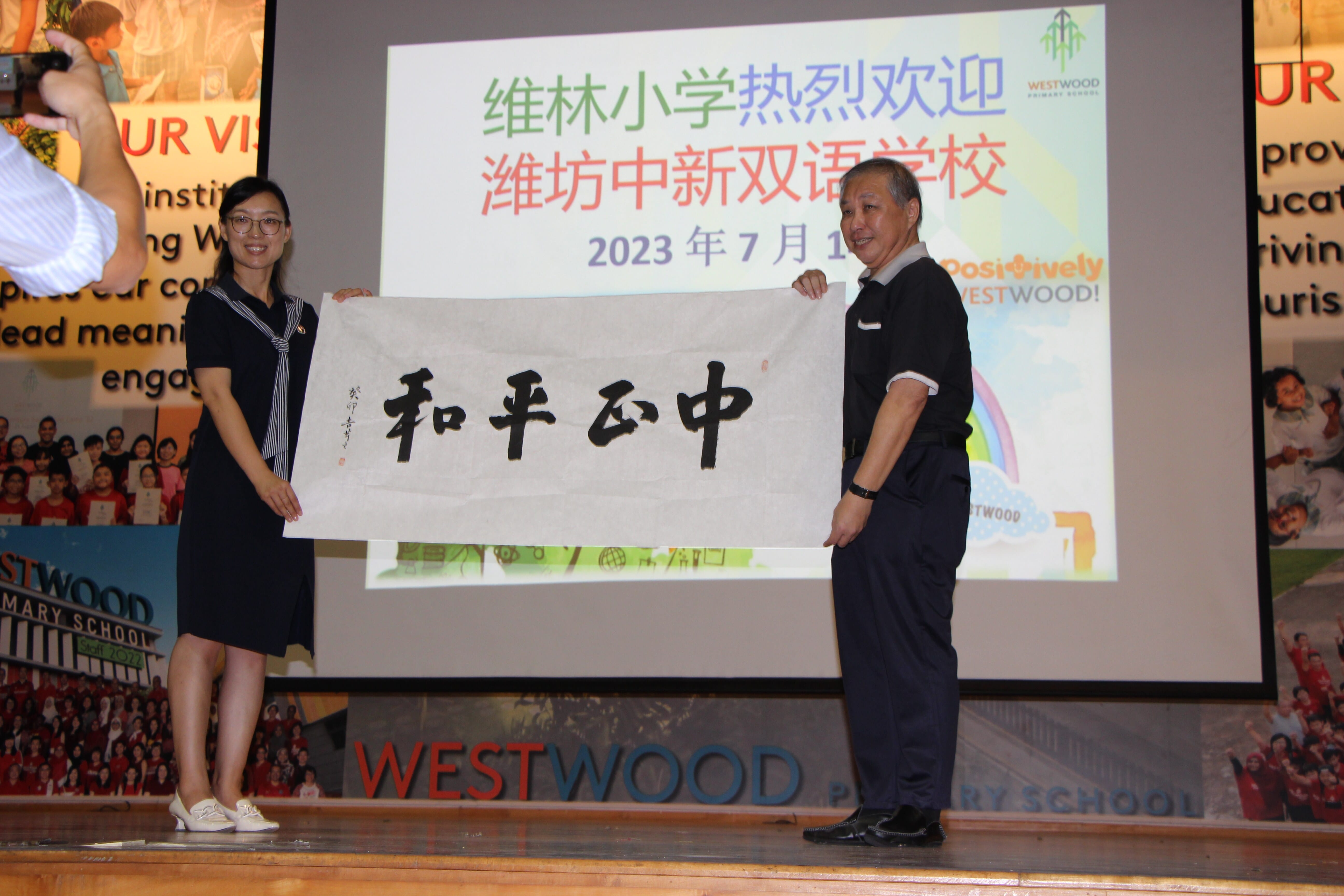 Two people stand on a stage at Westwood Primary School holding a scroll with Chinese calligraphy characters.